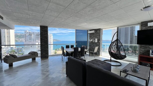 A spacious living room with a balcony overlooking a cityscape and the sea. - Apartment with ocean and golf course views (Acapulco de Juárez)