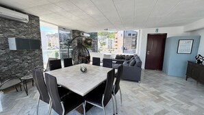 A modern living room with a dining area, stone wall, large windows, and a view of buildings. - Apartment with ocean and golf course views (Acapulco de Juárez)