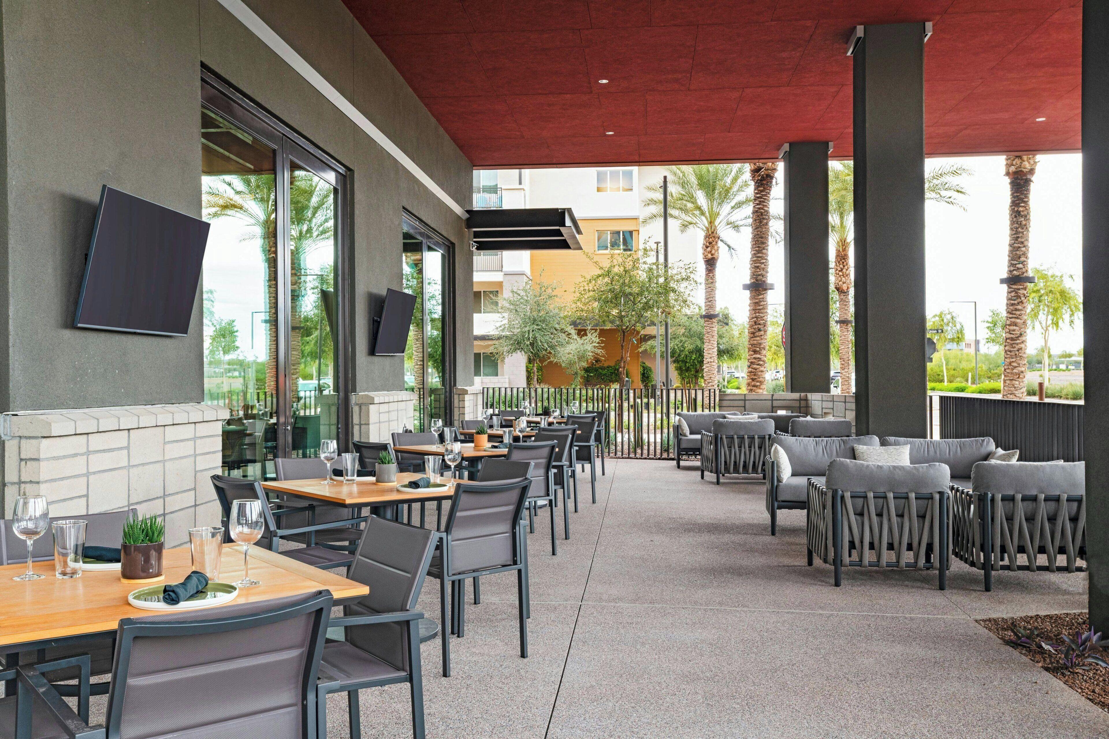 An outdoor dining area with tables and chairs, a flat-screen TV mounted on the wall, and a view of palm trees and buildings in the background.