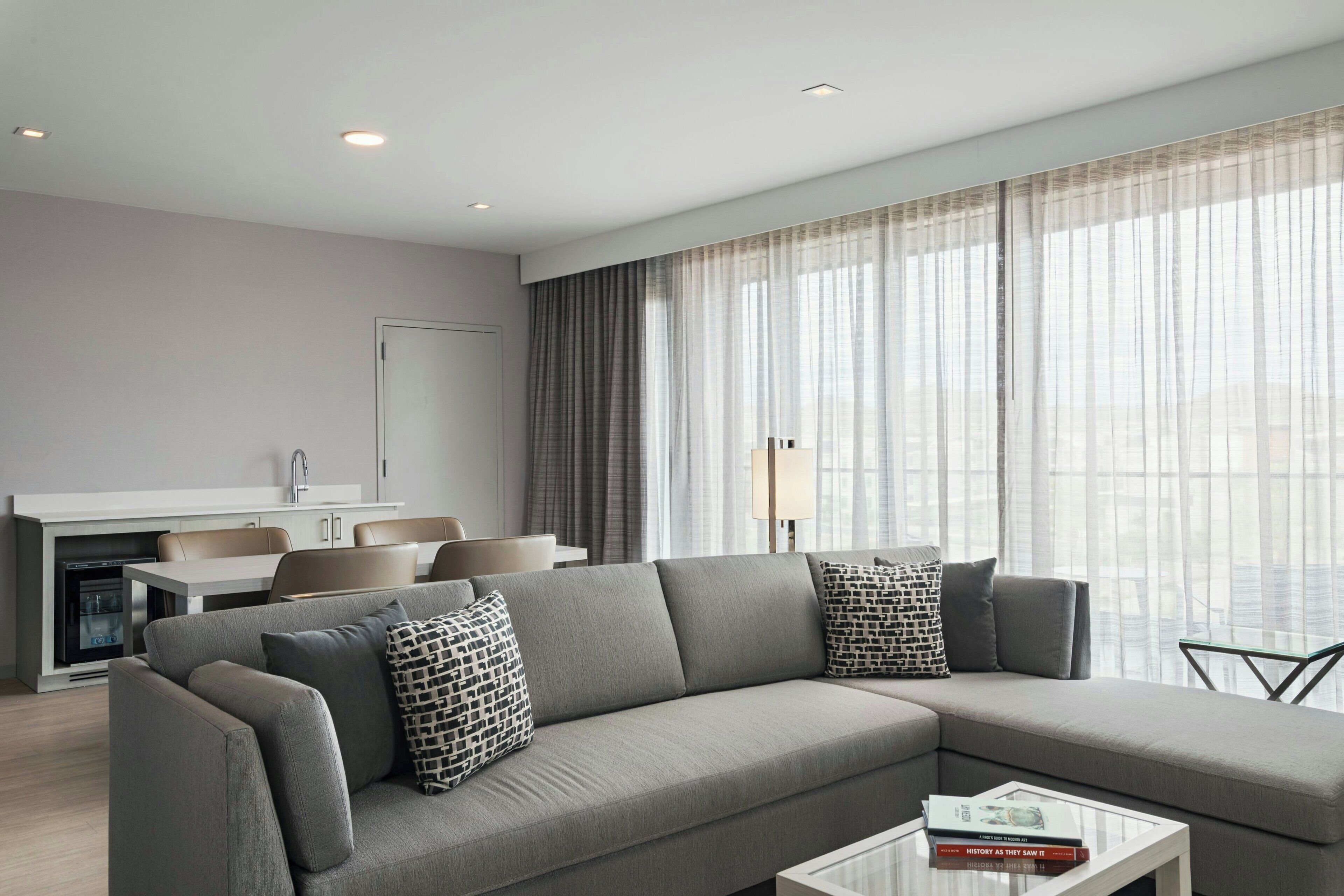 A modern living room with a grey sectional sofa, a glass coffee table, and a view of the city through large windows.