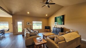 A spacious living room with a high ceiling, a ceiling fan, a large window with a view of the outdoors, a dining area with a table and chairs, and a television on a stand. - Experience Desert Gemini Ranch and the magic of the Colorado high desert (Fruita)