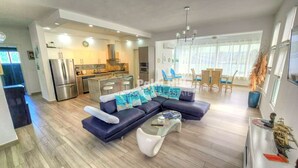 Un salon moderne avec un canapé d’angle bleu foncé, une table basse en verre et un coin repas doté de chaises en osier.