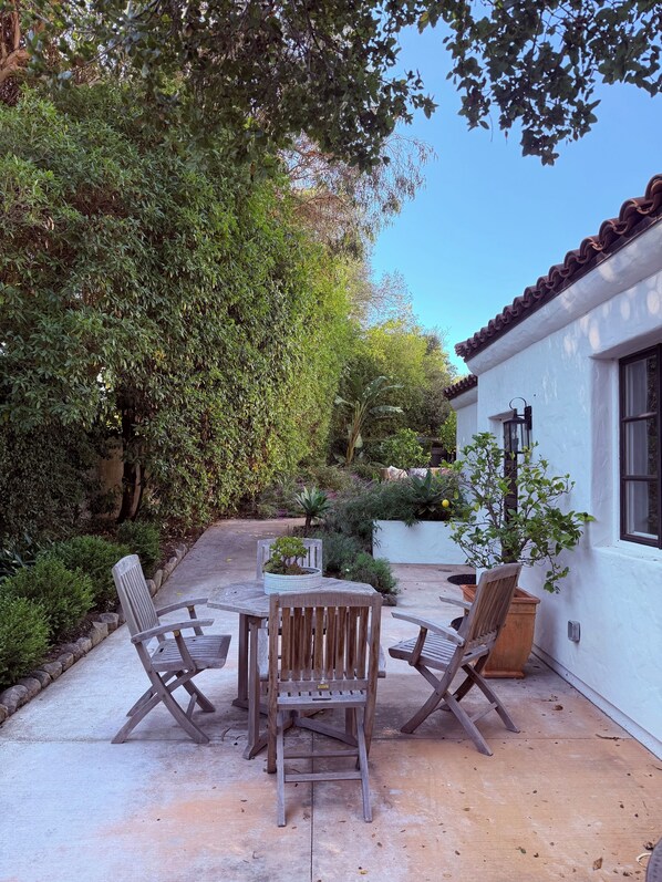 Un patio con una mesa y cuatro sillas de madera, rodeado de vegetación exuberante y un edificio blanco con techo de tejas.
