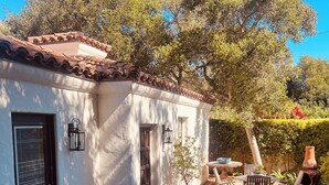A white building with a tiled roof, a door, and windows. There is a patio with a table and chairs, surrounded by plants and a large potted plant.