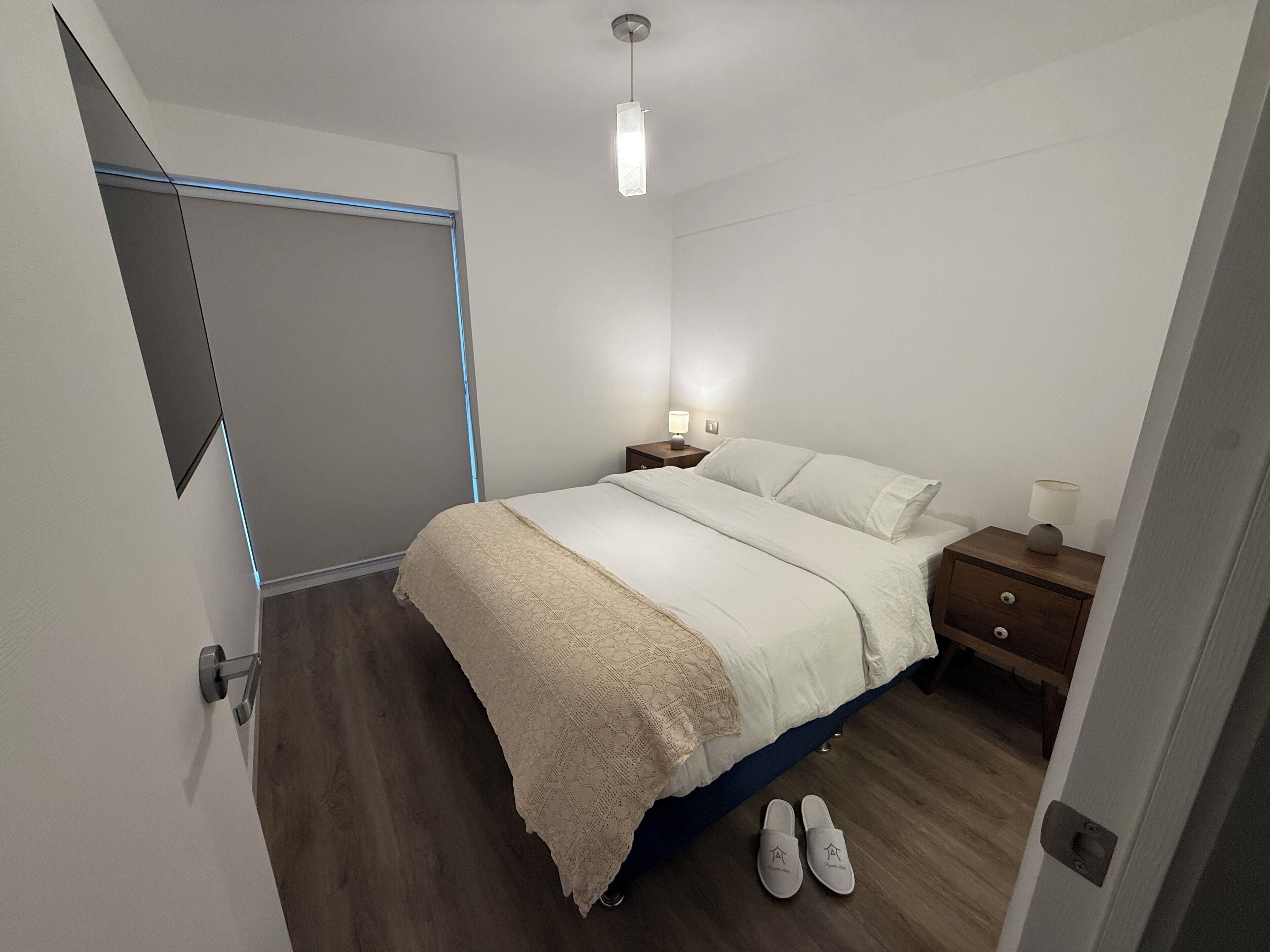 Quarto com cama, mesas de cabeceira, um criado-mudo, abajur e porta com puxador.