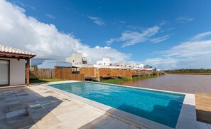 Un espace piscine moderne avec une piscine à l’eau d’un bleu limpide, une terrasse en bois et une vue sur un ensemble résidentiel composé d’immeubles blancs et beiges.