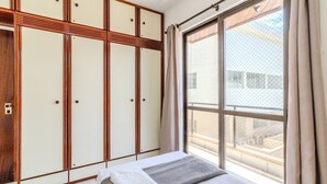 Ein Schlafzimmer mit großem Fenster, einem Bett mit grauer Decke und einem Schrank mit Holzeinlagen.