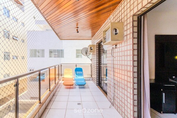 A balcony with two lounge chairs, a wooden ceiling, and a view of a building across a chain-link fence.