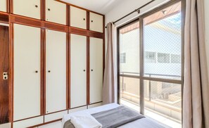 Une chambre avec une grande fenêtre, un lit recouvert d’une couverture grise et une armoire aux panneaux en bois.