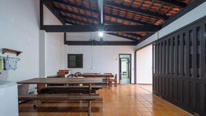 Amplia habitación con techo alto, vigas de madera y suelo de baldosas. Se observan varias mesas y bancos de madera dispuestos para comedor.