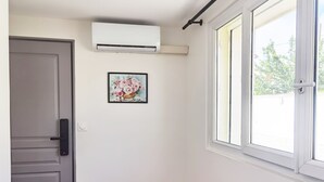 A room with a grey door, a white wall, a framed floral painting, an air conditioning unit, and a window with a view of greenery outside. - New studio with AC / free charging parking（NO.5） (Villemomble)