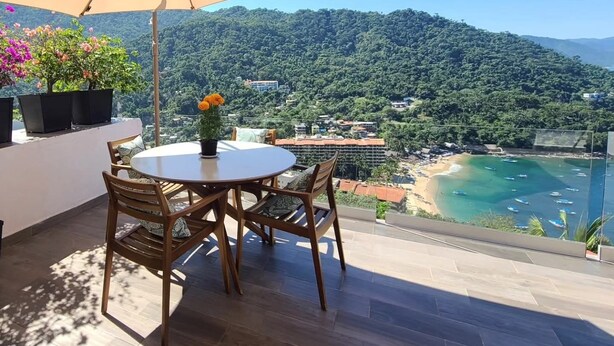 A terrace with a round table and chairs, a large umbrella, and a view of a coastal area with mountains in the background. - Charming 2-bedroom, 2 bath condo in Mismaloya with AC, remodeled kitchen & baths (Mismaloya)