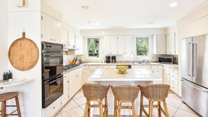 A modern kitchen with white cabinets, stainless steel appliances, a large island with bar stools, and a hanging wooden board. - Charming Sea Island Cottage • Steps to Beach • Sleeps 10 (Sea Island)