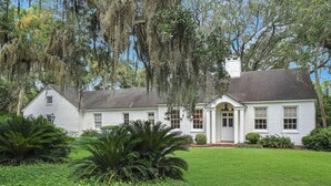 A white colonial-style house with a well-maintained lawn and large trees with hanging moss. - Charming Sea Island Cottage • Steps to Beach • Sleeps 10 (Sea Island)