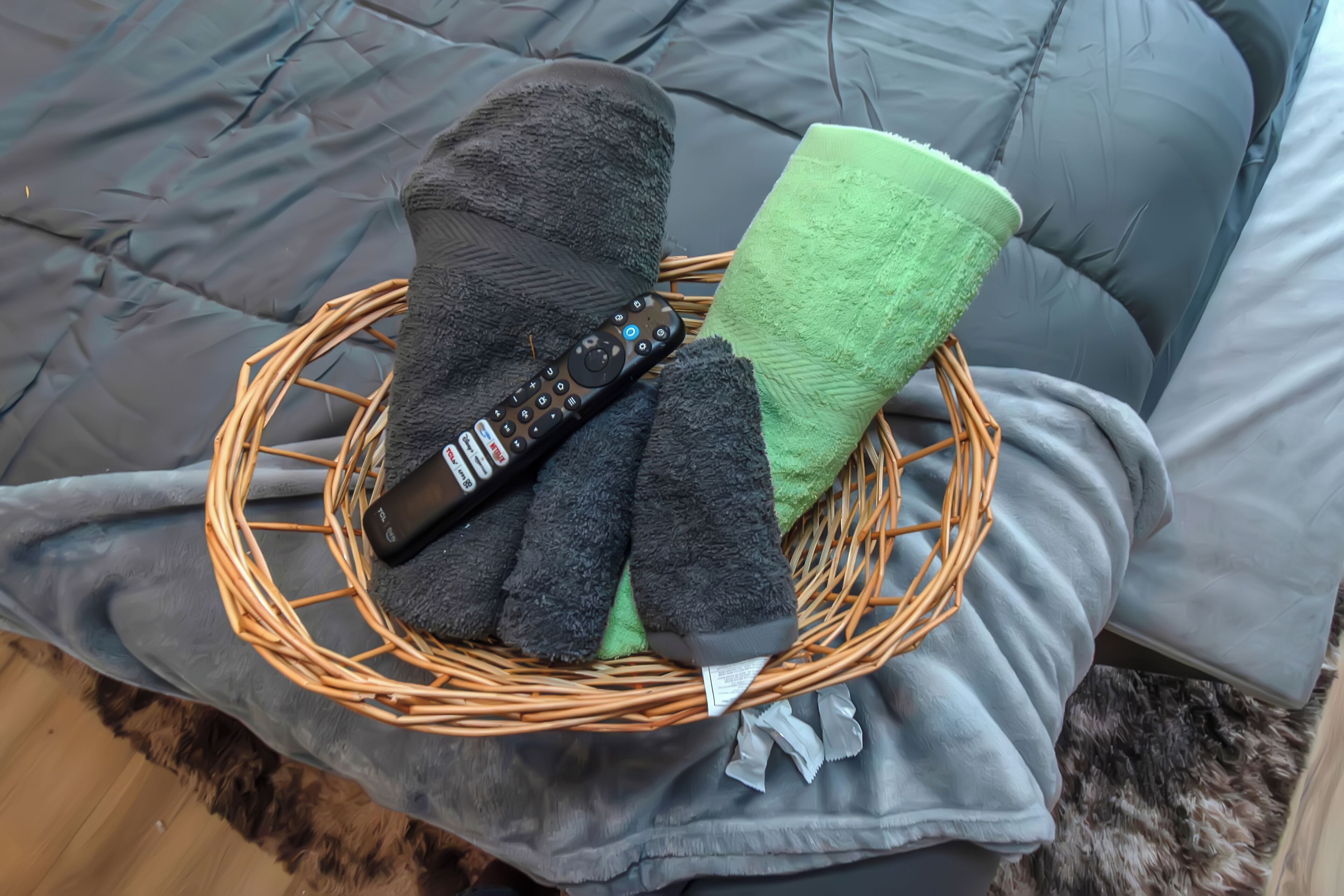 A wicker basket on a bed with a remote control, black and green slippers, and a grey blanket.