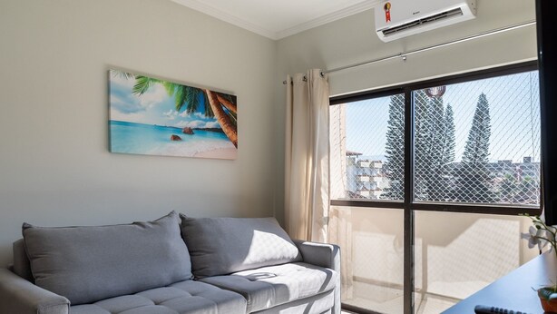 A modern living room with a grey sofa, a painting of a beach scene, and a large window with a view of buildings and trees. - Cozy Apartment Near the Beach Ubatuba Enu011 (Ubatuba)