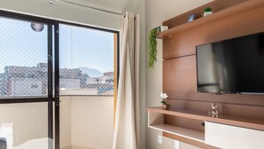 A modern living room with a large window, a flat-screen TV, wooden shelves, and a small plant. - Cozy Apartment Near the Beach Ubatuba Enu011 (Ubatuba)