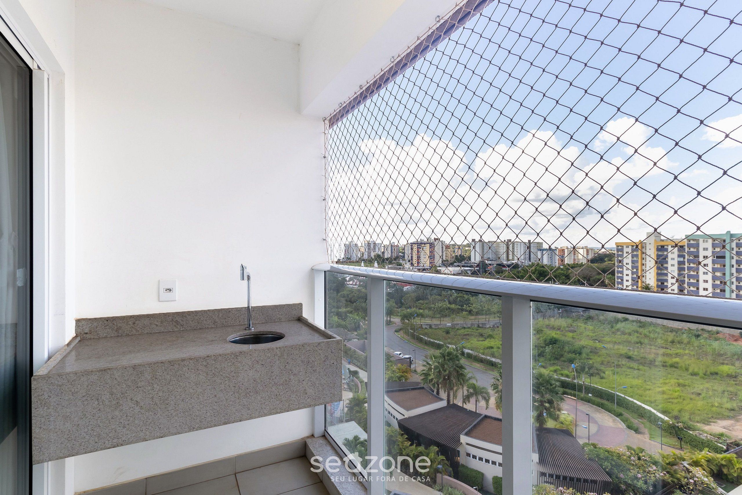A balcony with a metal safety net, a small sink, and a view of a cityscape with buildings and greenery.