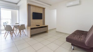 A modern living room with a flat-screen TV mounted on a wooden cabinet, a brown sofa, and a dining area with a table and chairs. - 3 Quartos no Evian Thermas Residence - Etha604 (Caldas Novas)