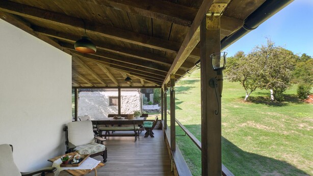 A covered patio area with wooden flooring, a table, chairs, and a view of a stone wall and grassy yard. - Cozy home in Petrinja with house a panoramic view (Petrinja)