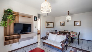 Una sala moderna con un televisor de pantalla plana, mueble de entretenimiento de madera, un área de comedor con mesa y sillas, y un candelabro.