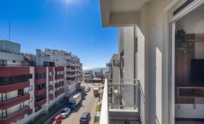 A high-rise building with multiple balconies, a clear view of the street below, and a distant body of water. - Apto 2Q 180m da praia com Garagem ERU032 (Bombinhas)