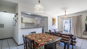 A dining area with a table covered in a floral tablecloth, wooden chairs, and a chandelier. - Apto 2Q 180m da praia com Garagem ERU032 (Bombinhas)