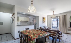 A dining area with a table covered in a floral tablecloth, wooden chairs, and a chandelier. - Apto 2Q 180m da praia com Garagem ERU032 (Bombinhas)