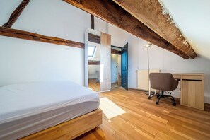 Quarto com cama de madeira, escrivaninha e cadeira. Há um claraboia e uma porta que dá para outro cômodo.