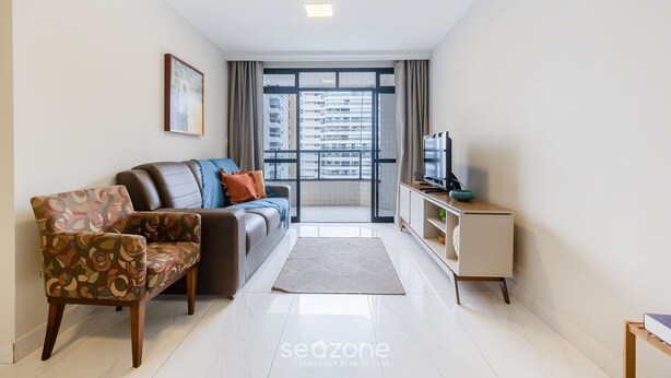 A modern living room with a sofa, a patterned armchair, a TV, and a view of a cityscape through large windows. - 3 Bedroom Apartment 190m From the Beach Epp0602 (Guarapari)