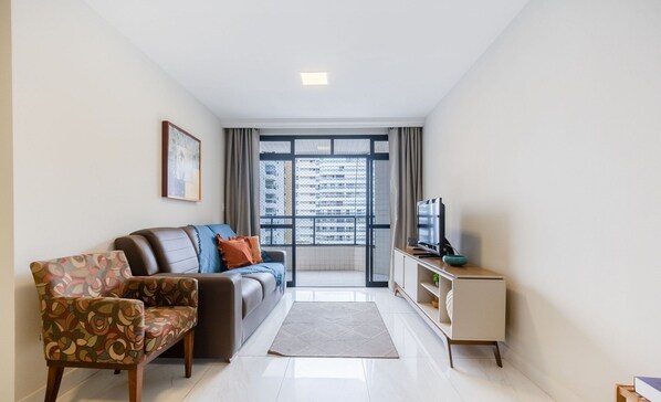 Una sala de estar moderna con un sofá, un sillón estampado, un televisor y vistas a un paisaje urbano a través de amplios ventanales.