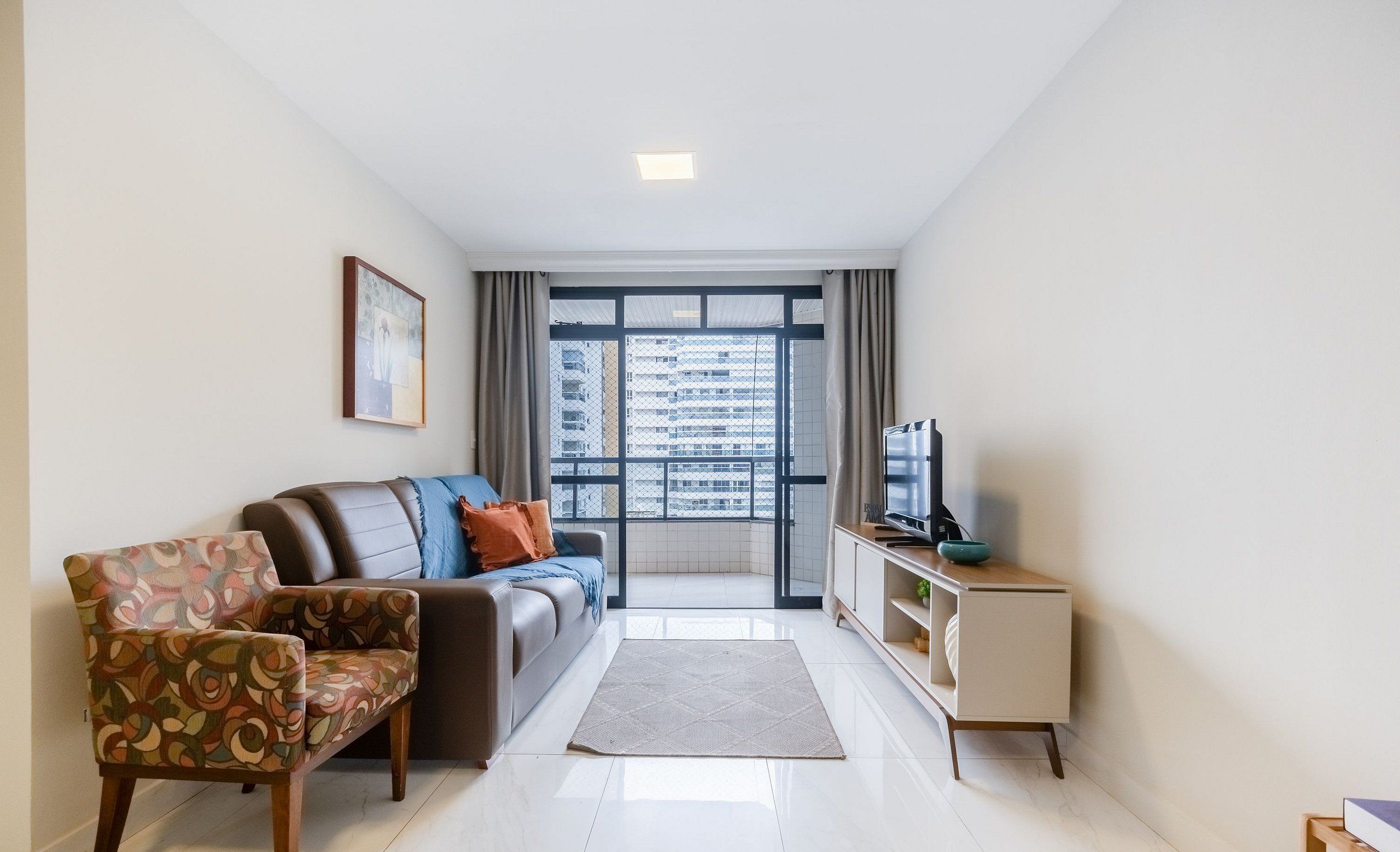 Uma sala de estar moderna com um sofá, uma poltrona estampada, uma TV e vista para um horizonte urbano por meio de grandes janelas.