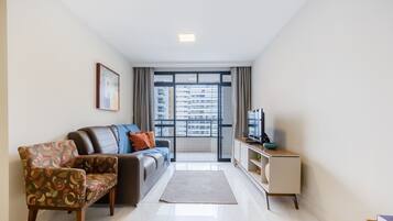 Uma sala de estar moderna com um sofá, uma poltrona estampada, uma TV e vista para um horizonte urbano por meio de grandes janelas.