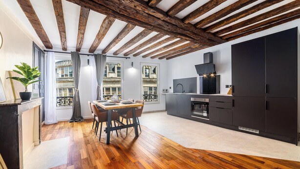 A modern kitchen with dark cabinetry, a wooden dining table, and a view of the city through large windows. - Triplex Belle Époque Hypercenter (Melun)