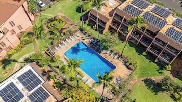 An aerial view of a hotel with a swimming pool surrounded by lounge chairs and palm trees, solar panels on the roof, and a well-maintained lawn.