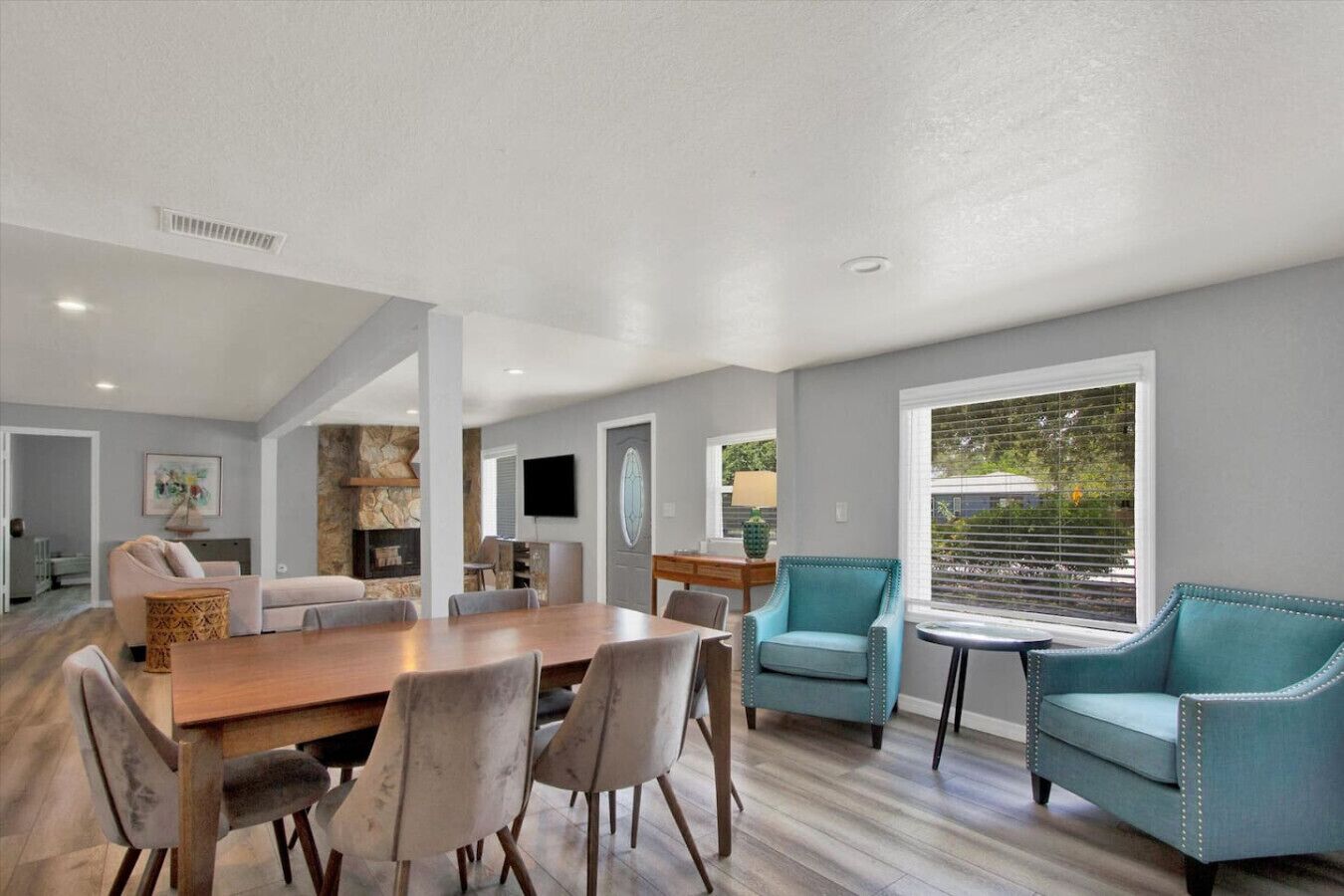 A spacious living room with a wooden dining table, comfortable seating, a stone accent wall, and a large window with blinds.
