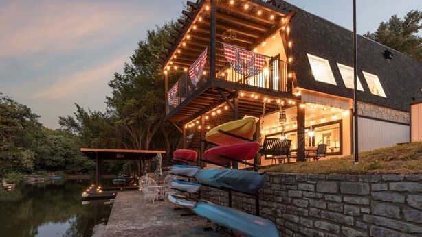 A two-story house with a deck overlooking a body of water, kayaks, and a stone wall.