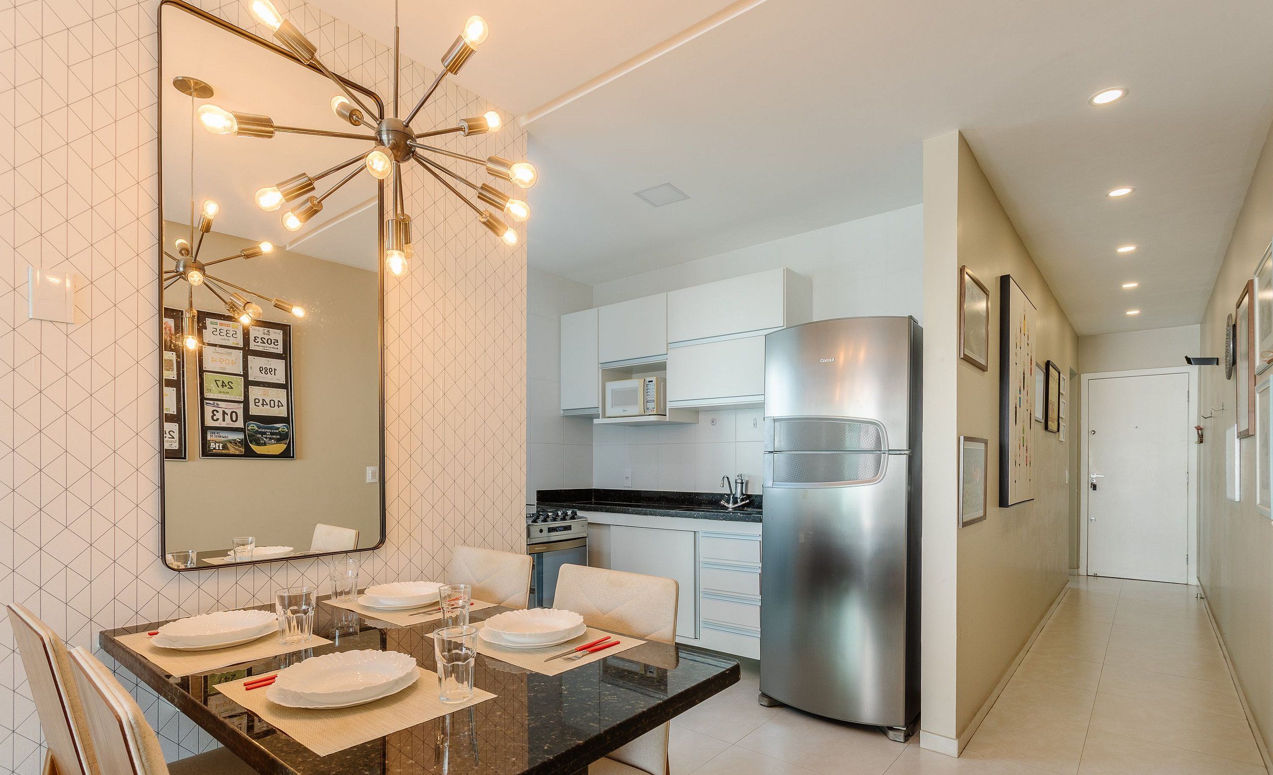 Una cocina moderna con armarios blancos, un refrigerador de acero inoxidable y un comedor con un espejo grande y luces colgantes.