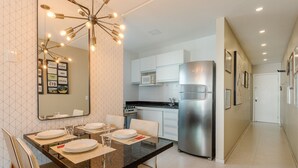 A modern kitchen with white cabinets, a stainless steel refrigerator, and a dining area with a large mirror and pendant lights. - Charming Apt Just 450m From the Beach Emm0202 (Guarapari)