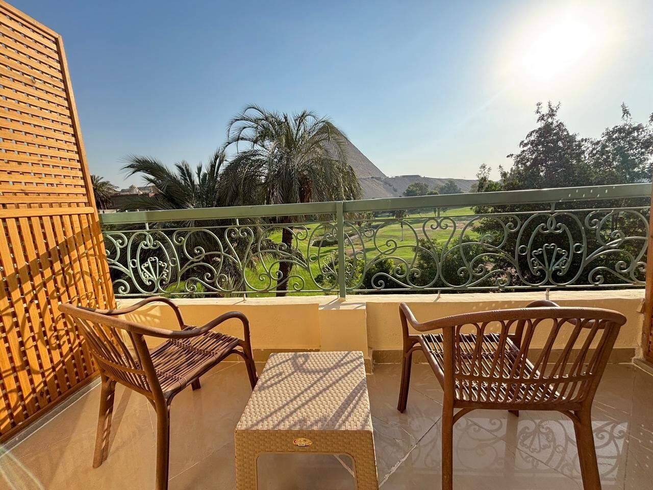 Un balcone con due sedie in vimini e un tavolino, affacciato su un paesaggio verde con palme e una piramide all'orizzonte.