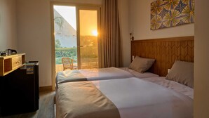 Habitación de hotel con dos camas, cabecero de madera, un escritorio pequeño con refrigerador y vistas al exterior a través de una puerta corrediza.