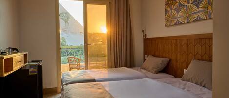 A hotel room with two beds, a wooden headboard, a small desk with a refrigerator, and a view of the outdoors through a sliding door.