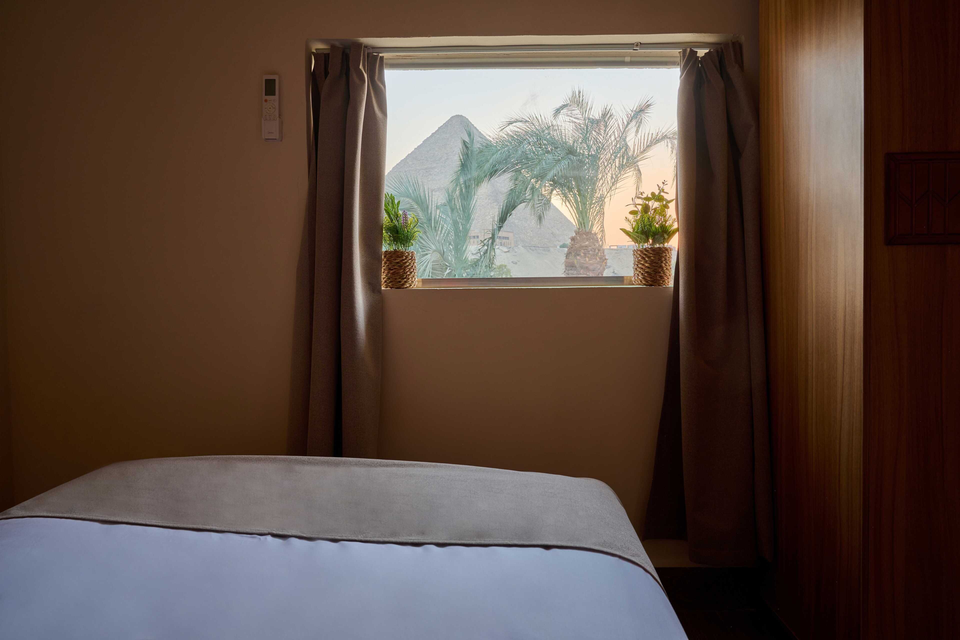 A hotel room with a bed, a window with curtains, and two potted plants on the windowsill.