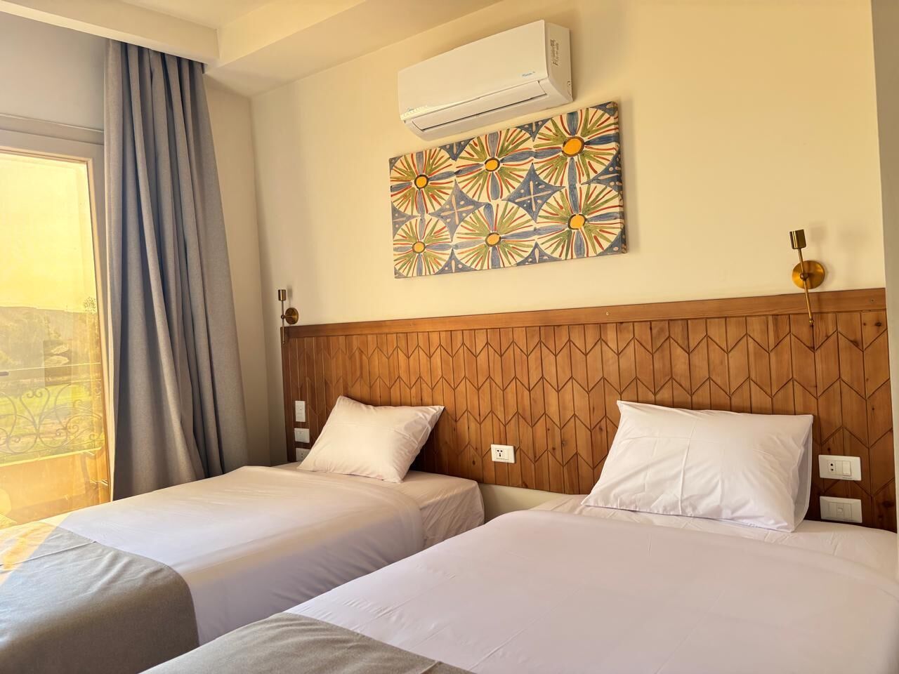A hotel room with two beds, a wooden headboard, an air conditioner, a patterned artwork, and a window with curtains.