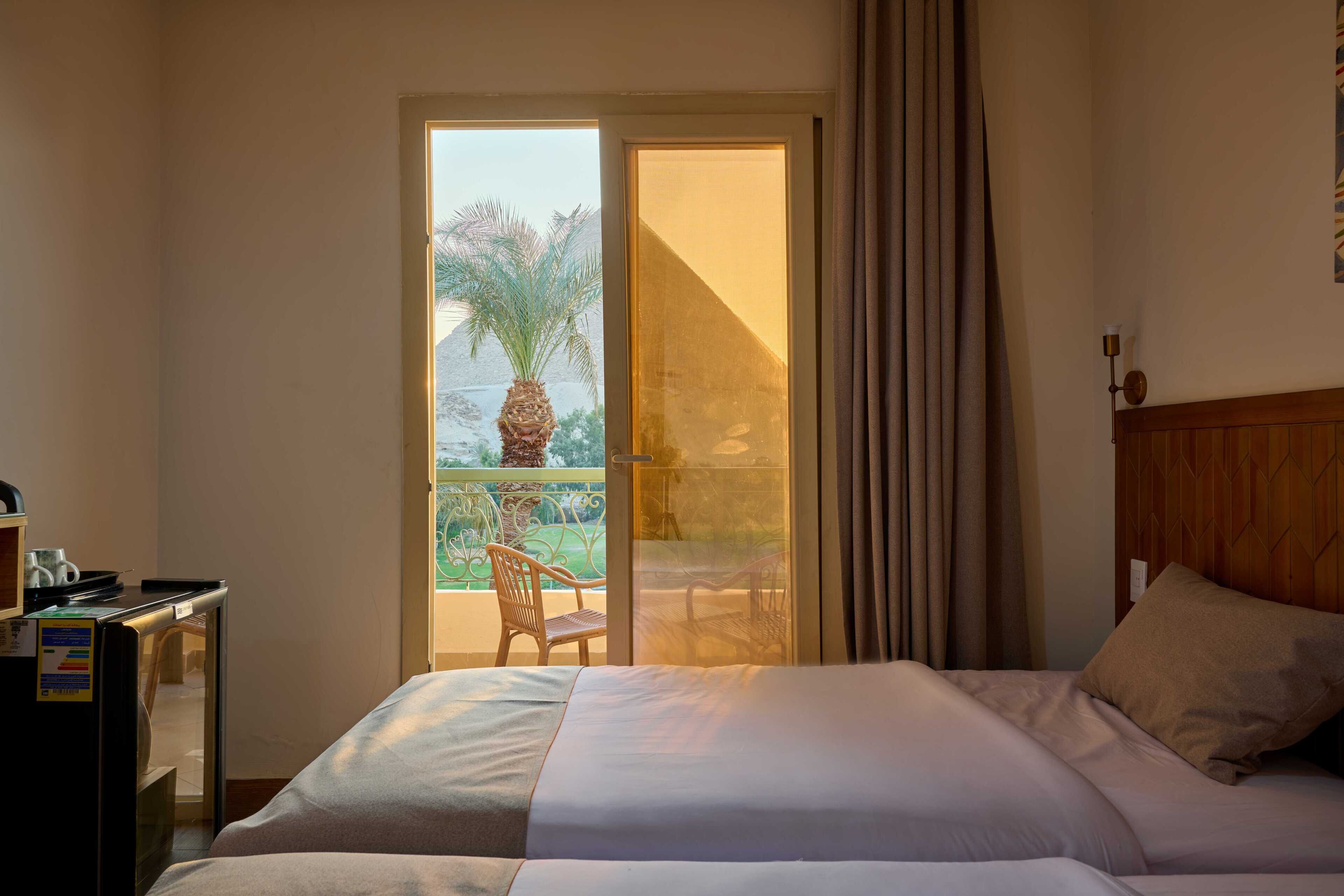 Quarto de hotel com cama, escrivaninha, cadeira e varanda com vista para palmeiras e montanhas.