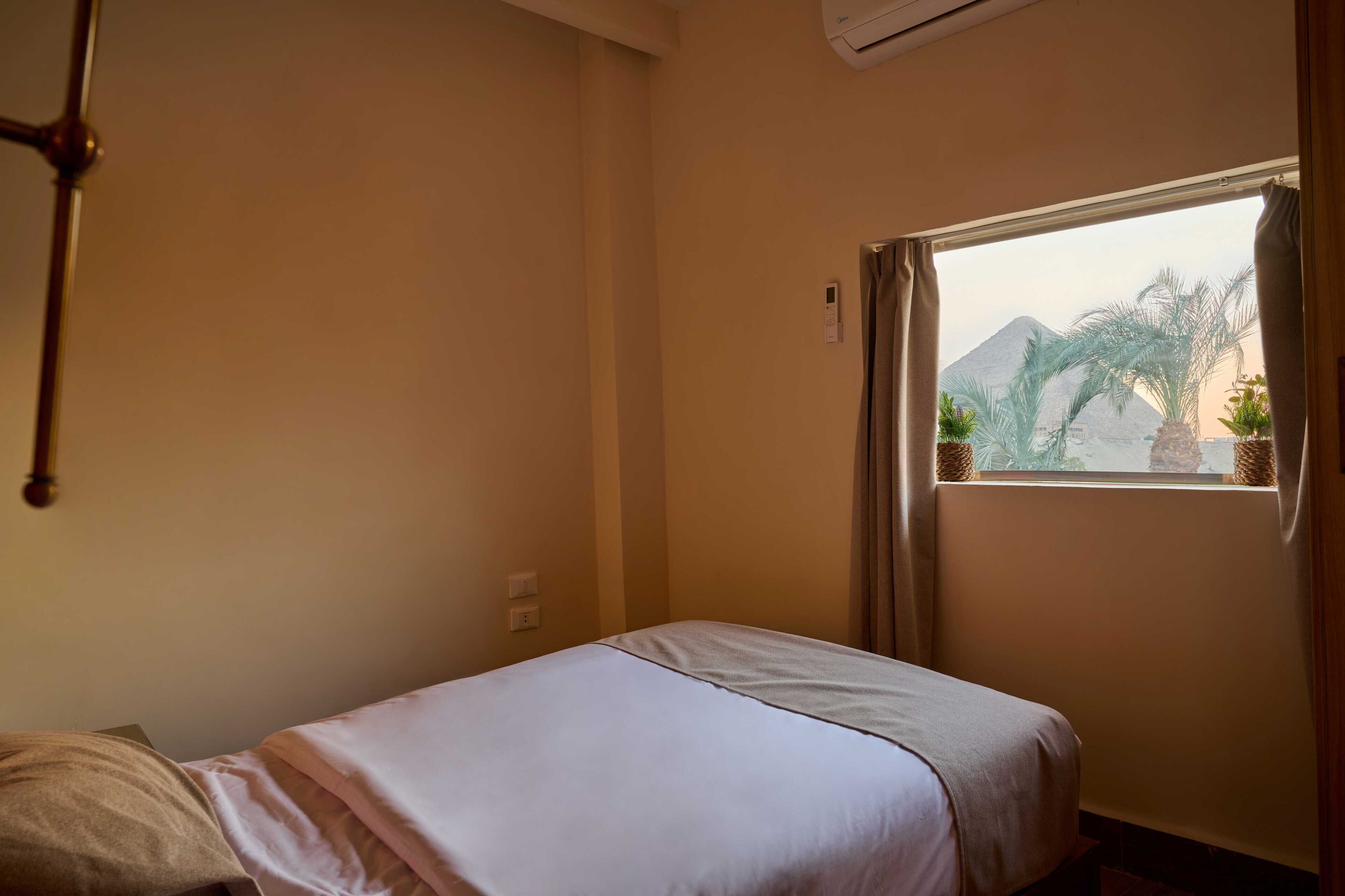 A single bed with a white bedspread in a room with beige walls, a window with curtains, and a view of palm trees and a mountain.