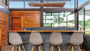 A bar area with a wooden counter, three bar stools, and a large window with a view of greenery and buildings. - 3BR Duplex w/ BBQ & Garage Near Beach Emb686 (Bombinhas)