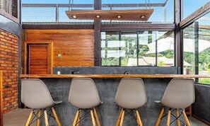 A bar area with a wooden counter, three bar stools, and a large window with a view of greenery and buildings. - 3BR Duplex w/ BBQ & Garage Near Beach Emb686 (Bombinhas)