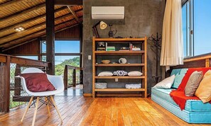 Ein Zimmer mit Holzbalkendecke, einem Bücherregal, einem weißen Stuhl, einem blauen Sofa und einem Fenster mit Vorhängen.