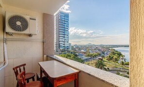 A balcony with a table and chairs, a wall-mounted air conditioner, and a view of a cityscape and water body. - Stay With an Ocean View! Elg0071 (Barra Velha)